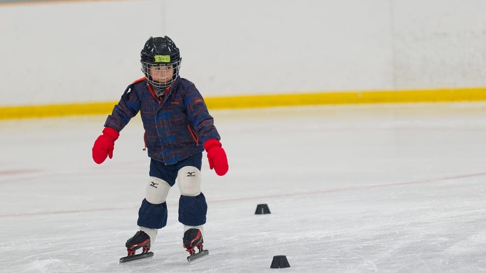 A young Learn to Skate kid in a hockey helmet and red mittens, mid-stride.