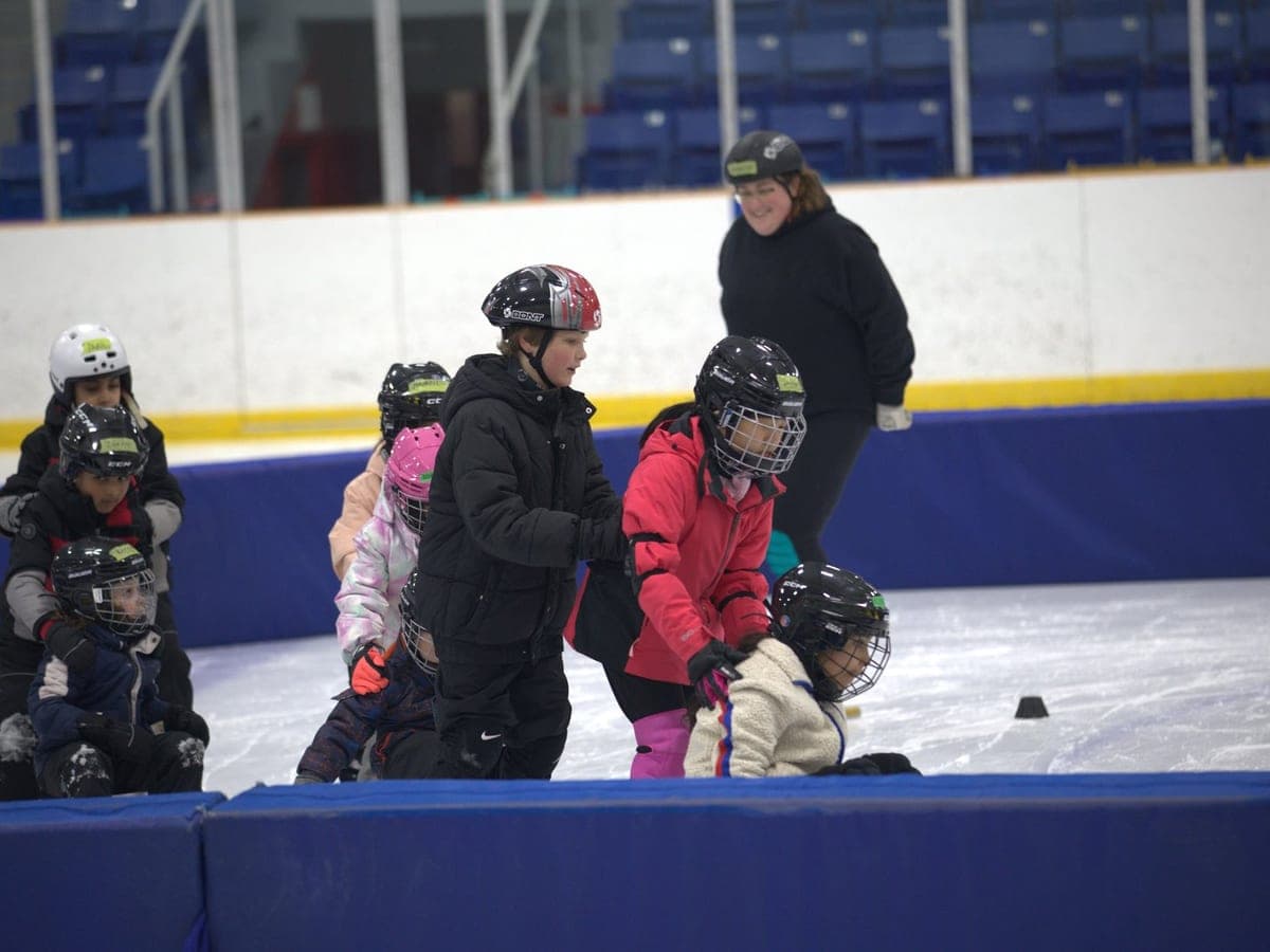 A group of Learn to Skate kids gathered near the boards with a coach behind them.