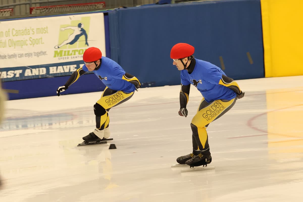 Two Masters skaters in matching Golden Horseshoe kit, side-by-side mid-stride on the ice.